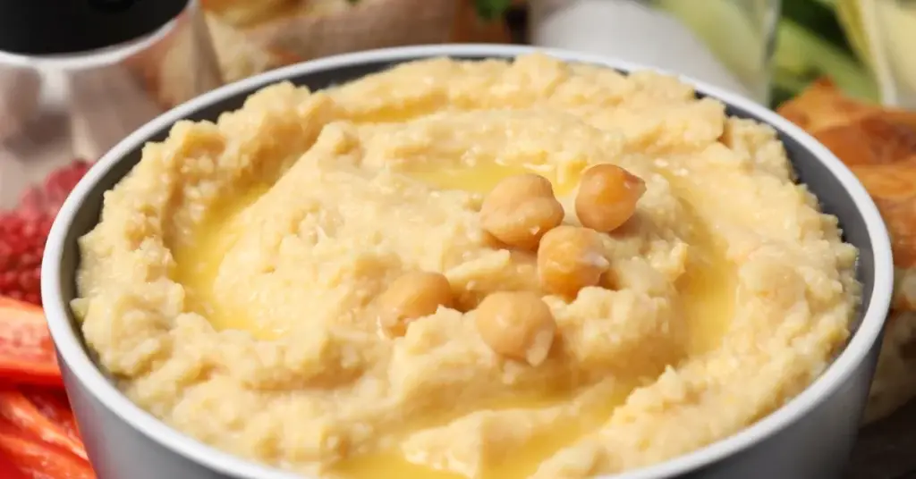 Creamy hummus topped with chickpeas and a drizzle of golden rapeseed oil, served in a bowl with fresh vegetables nearby.