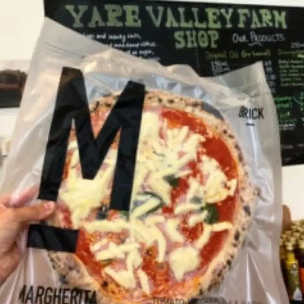 Brick Margherita pizza held inside Yare Valley Farm Shop with the shop chalkboard visible in the background.