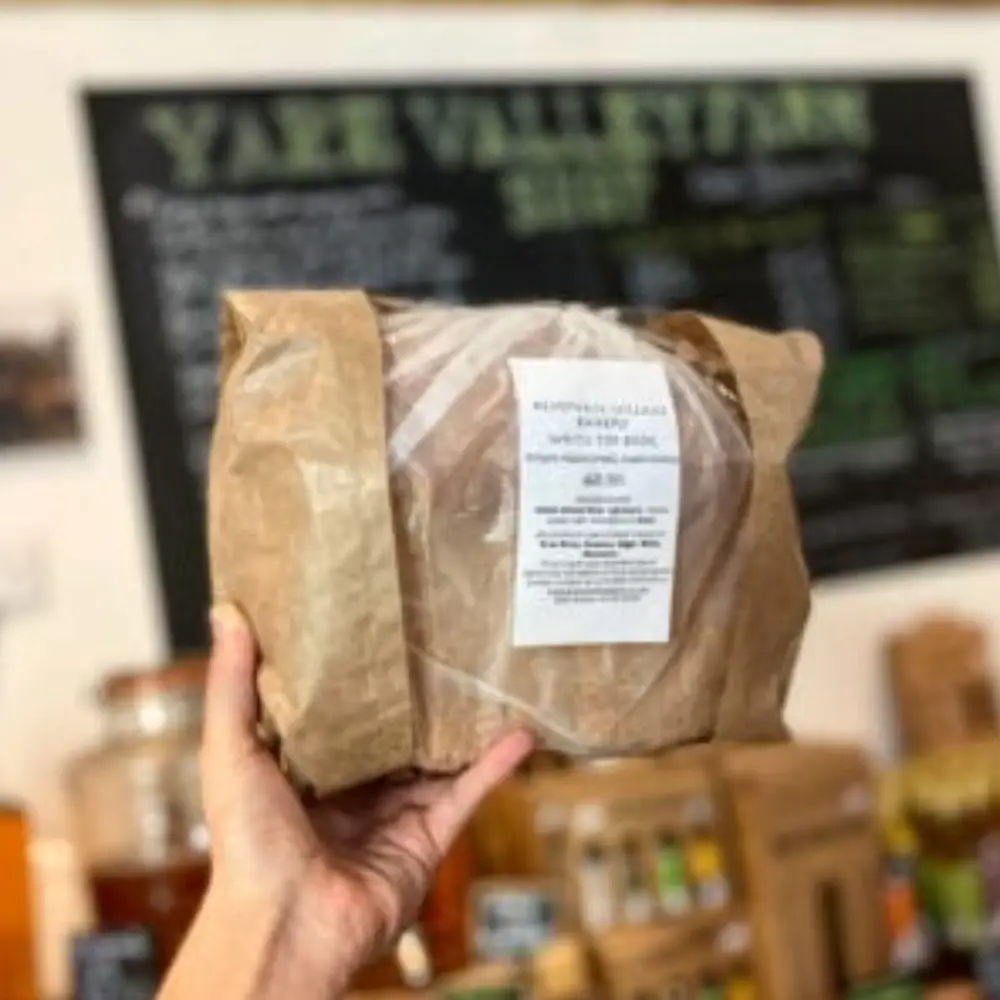 Wrapped loaf of sourdough bread held inside Yare Valley Farm Shop with the shop chalkboard in the background.