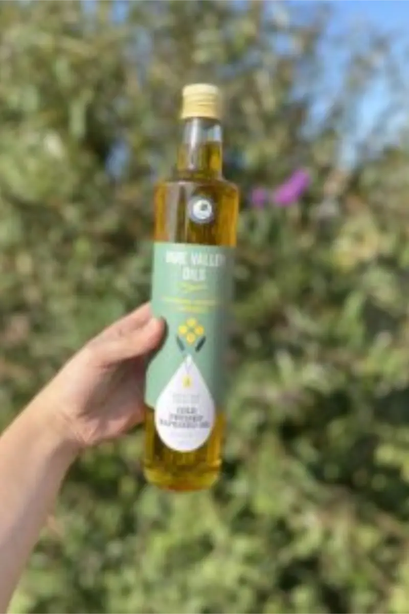 Hand holding a tall bottle of Yare Valley rapeseed oil in front of trees and wild greenery.